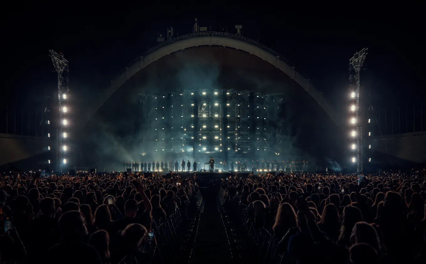 A concert with a large crowd in front of a brightly lit stage at night, featuring a performer and a line of singers in the background.