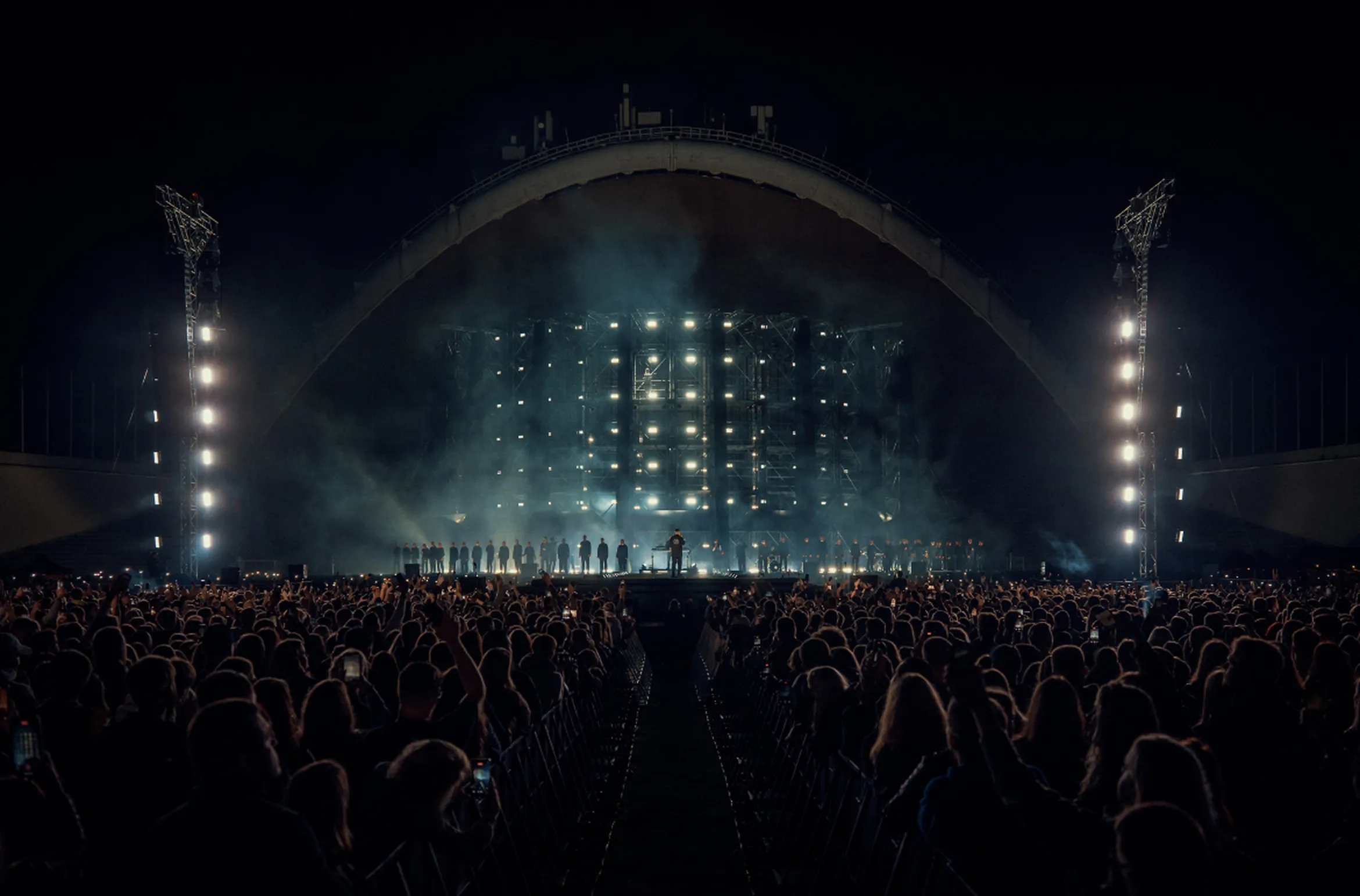 A concert with a large crowd in front of a brightly lit stage at night, featuring a performer and a line of singers in the background.