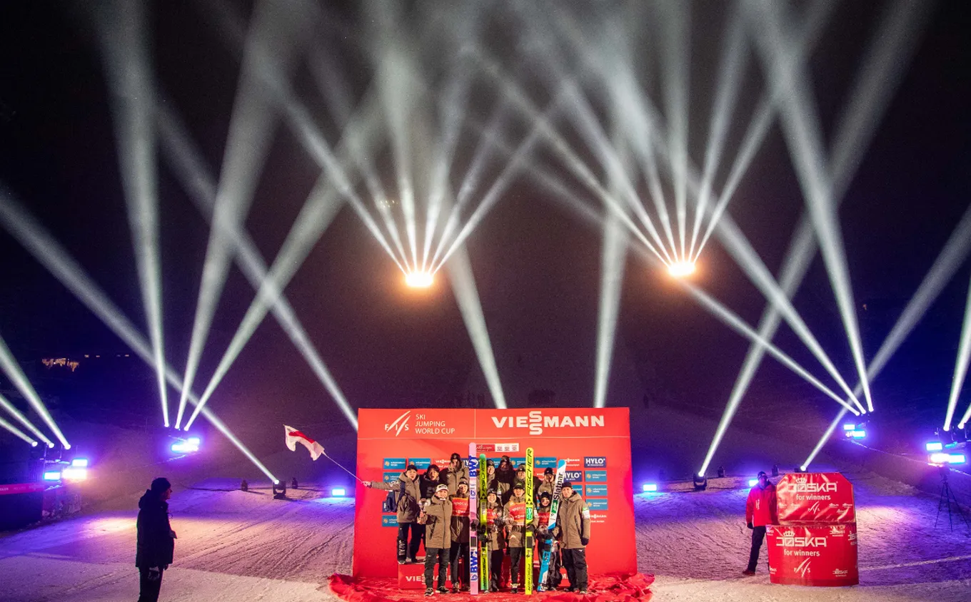 Skiing World Cup podium ceremony with winners holding skis, surrounded by bright spotlights and a red backdrop in a snowy night setting.