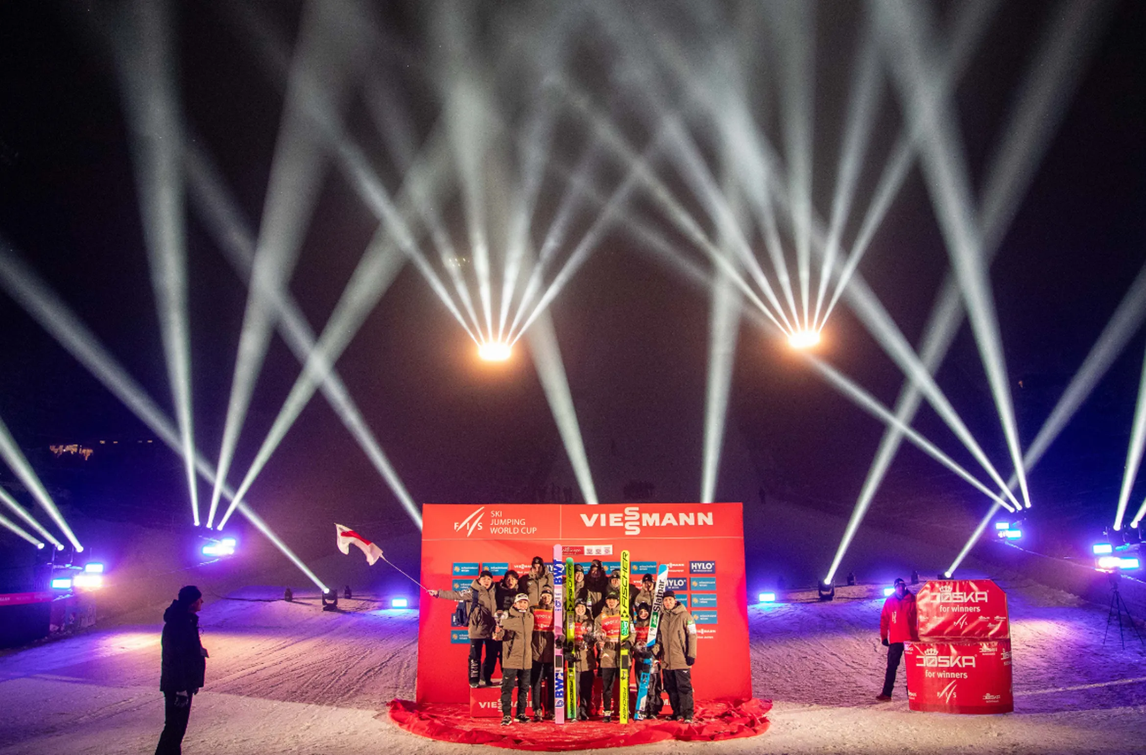 Skiing World Cup podium ceremony with winners holding skis, surrounded by bright spotlights and a red backdrop in a snowy night setting.